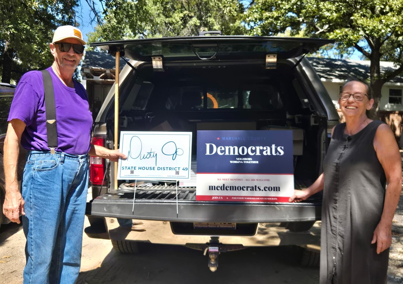 Two Marshall County Democrats volunteers holding yard signs at the tailgate of a truck — a navy Marshall County Democrats sign reading mcdemocrats.com and a Dusty Cline for State House District 49 sign.